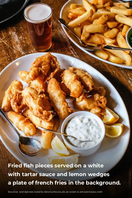 Fried fish pieces served on a white plate with tartar sauce and lemon wedges with a plate of french fries in the background.