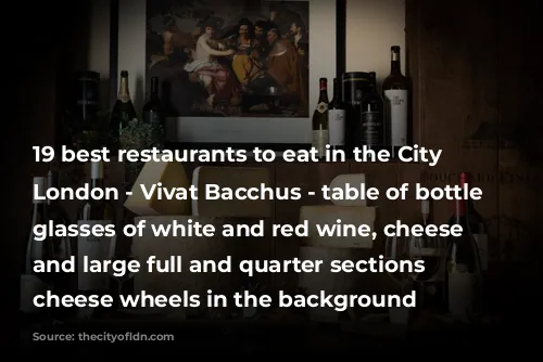 19 best restaurants to eat in the City of London - Vivat Bacchus - table of bottle and glasses of white and red wine, cheese boards and large full and quarter sections of cheese wheels in the background