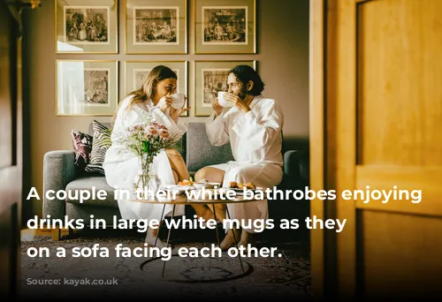 A couple in their white bathrobes enjoying their drinks in large white mugs as they sit on a sofa facing each other.