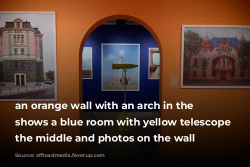 an orange wall with an arch in the centre shows a blue room with yellow telescope in the middle and photos on the wall