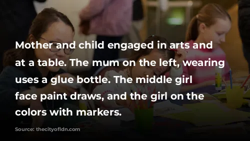 Mother and child engaged in arts and crafts at a table. The mum on the left, wearing glasses, uses a glue bottle. The middle girl with face paint draws, and the girl on the right colors with markers.