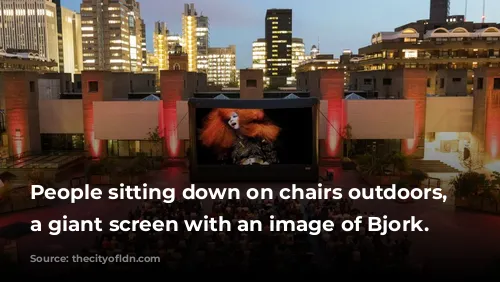 People sitting down on chairs outdoors, watching a giant screen with an image of Bjork.