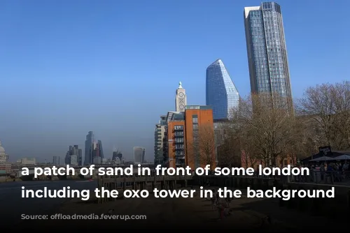 a patch of sand in front of some london buildings, including the oxo tower in the background